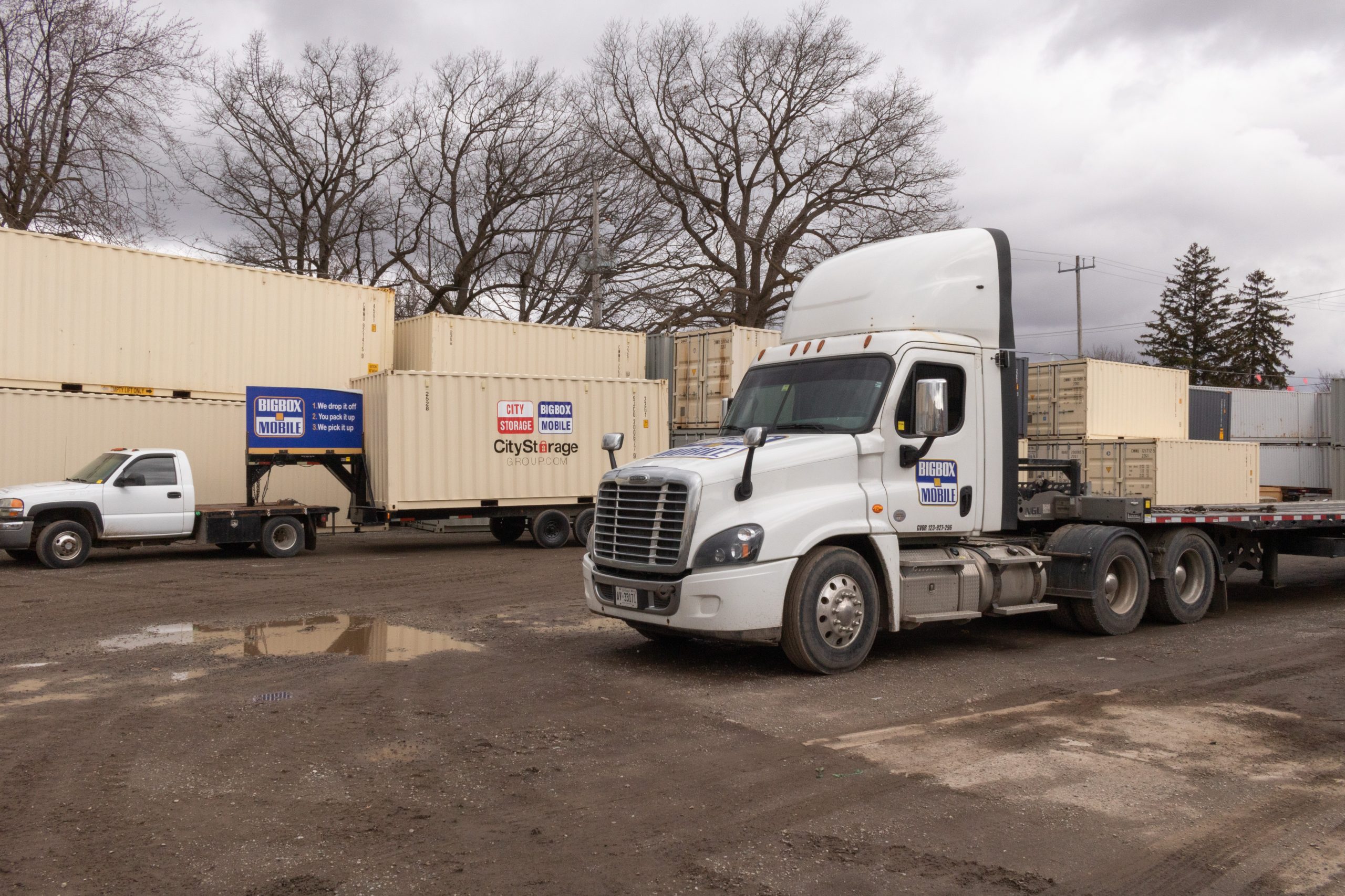 different trucks used to deliver mobile storage units in front of stacked shipping container storage bins